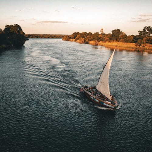 Ein Segelboot mit weißem Segel segelt auf einem breiten Fluss, umgeben von Grünland bei Sonnenuntergang.