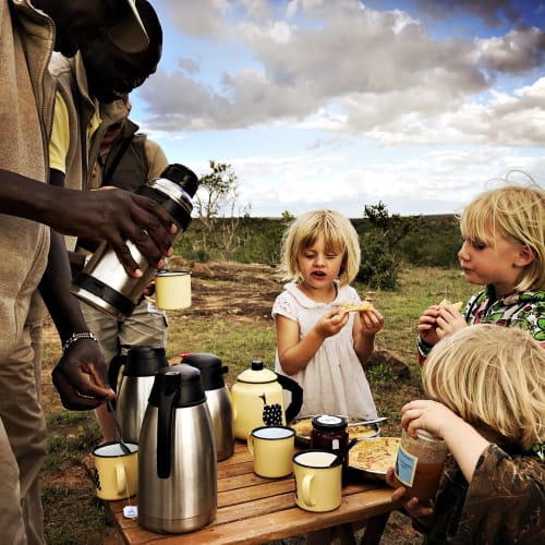 Three young children and an adult enjoying breakfast outdoors at a rustic wooden table in a natural setting with cloudy sky.
