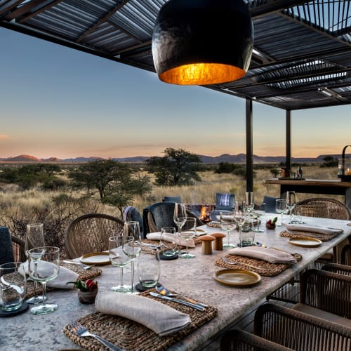 An outdoor dining table set on a deck overlooking the Kalahari desert at sunset, with chairs and tableware in place.