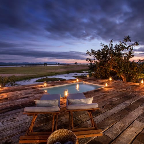 A cozy veranda overlooking a river with candles at twilight, featuring wooden furniture and a large tree nearby.
