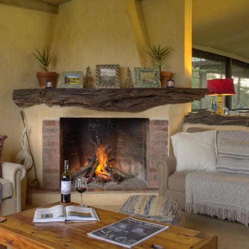 Gemütliche Lounge mit einem brennenden Kamin, umgeben von Sessel und einem Holztisch, in einem rustikalen Ambiente.