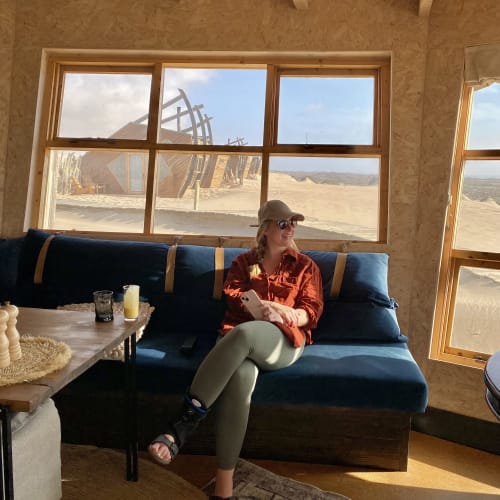 Eine Frau mit beiger Mütze, Sonnenbrille und orangenem Hemd sitzt mit übereinandergeschlagenen Beinen auf einer blau gepolsterten Bank in einer Holzhütte, hält ein Smartphone und schaut aus dem Fenster auf eine sandige Wüstenlandschaft mit einzigartigen Holzstrukturen draußen.