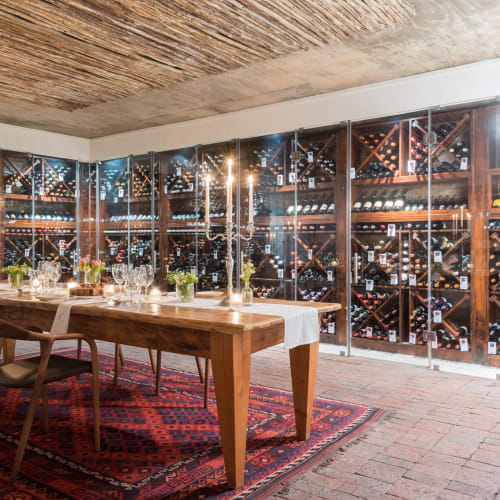 Ein gemütlicher Weinkeller mit Essbereich, Holztischen, Stühlen und hohen brennenden Kerzen, umgeben von Glasschränken mit zahlreichen Weinflaschen, unter einer rustikalen Decke mit einem gemusterten Teppich auf dem Boden.