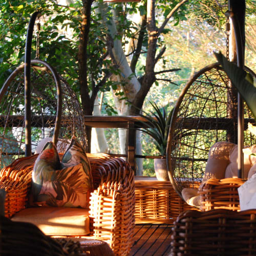 A cozy veranda lounge with wicker chairs and hanging egg chairs surrounded by lush greenery and sunlight filtering through the trees.