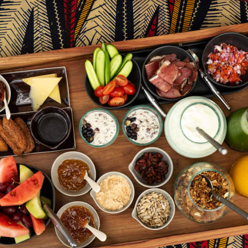 Assorted breakfast foods including fresh fruits, vegetables, yogurt, bread, and toppings on a wooden tray with a patterned fabric background.