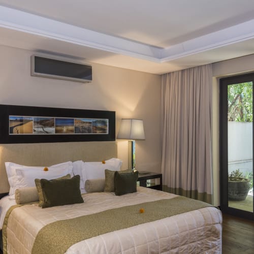 A well-lit bedroom with a large bed, multiple pillows, two bedside lamps, and a sliding glass door leading to an outdoor garden area.