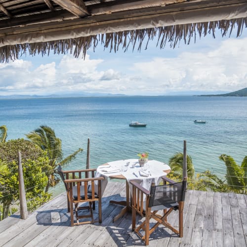 Ein Holzdeck mit einem für eine Mahlzeit gedeckten Tisch mit Blick auf den Ozean, Boote im Wasser und üppige tropische Pflanzen am Rand des Decks.