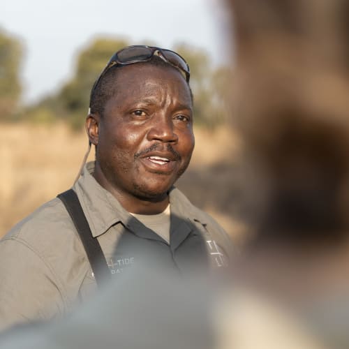 Ein Safariguide im Freien mit Bäumen und klarem Himmel im Hintergrund, trägt ein beiges Hemd und Sonnenbrille auf dem Kopf.