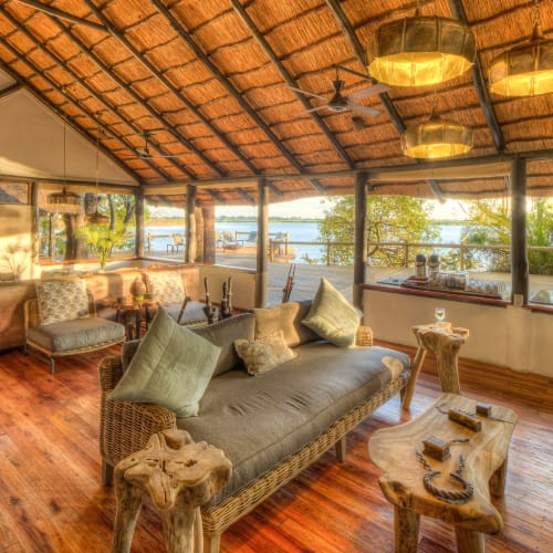 A spacious lounge and bar area with large windows overlooking a water body and surrounding greenery, featuring wooden floors, a sofa with pillows, and a wooden ceiling.