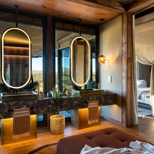 Luxurious bathroom with two illuminated mirrors, large windows showing nature, and a seating area nearby in a modern, elegant setting.