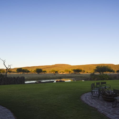 Ein geräumiger Außenbereich mit Stühlen und offenem Feuer in der Nähe einer üppigen grünen Wiese vor goldenen Hügeln in Namibia.