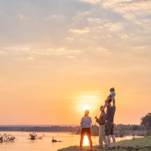 Eine Familie genießt einen Sundowner am Fluss bei Sonnenuntergang in South Luangwa, mit tiefstehender Sonne am Himmel und wenigen Wolken.
