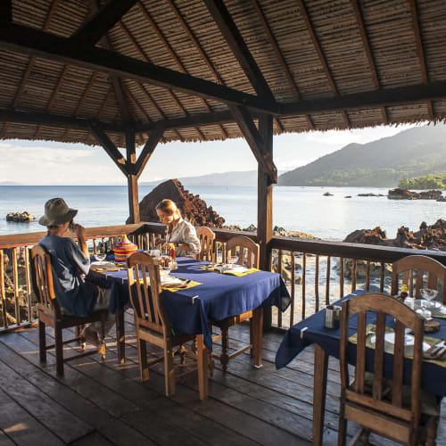 Breakfast setup on a shaded wooden terrace overlooking the ocean and rugged coastline.