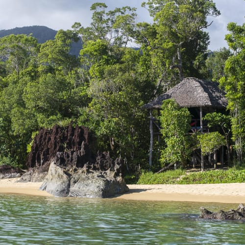 Ein Strand mit einer strohgedeckten Hütte, umgeben von dichtem grünen Wald am Ufer eines ruhigen Gewässers in Madagaskar.