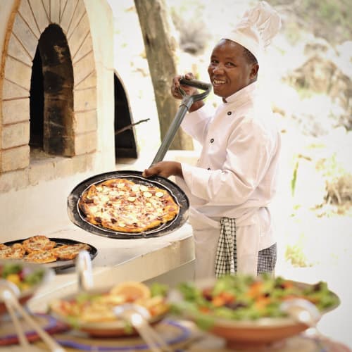 A smiling chef wearing a white uniform and hat lifts a freshly baked pizza out of a traditional outdoor oven, with a table of salads in the foreground.