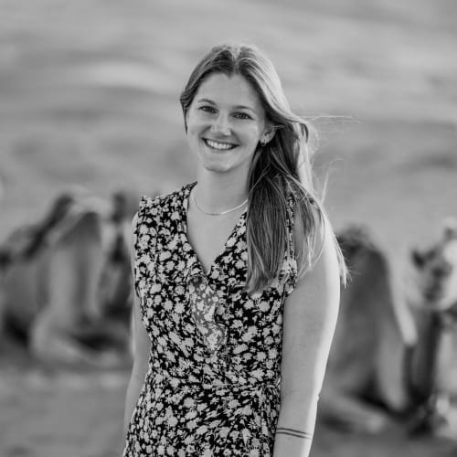 A smiling young woman with long hair stands outdoors with lions resting in the background.