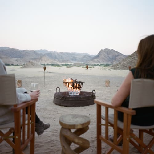 Zwei Personen sitzen bei einem Lagerfeuer in einer Wüstenlandschaft und genießen Getränke in Namibia.