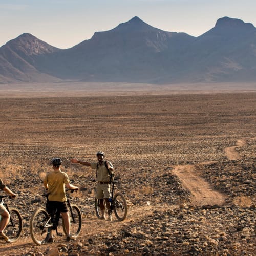 Three cyclists ride their bikes through a vast, arid desert landscape with mountains in the distance.