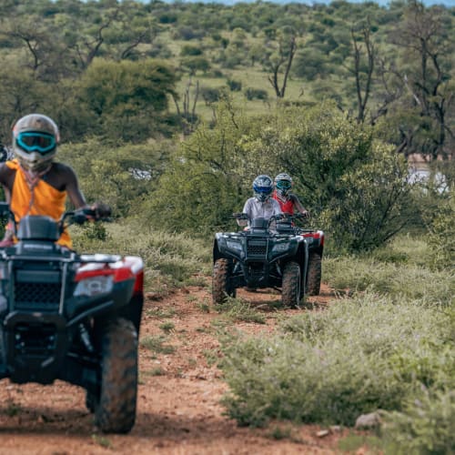 Three people riding all-terrain vehicles on a dirt trail through a semi-arid landscape with bushes and sparse trees.
