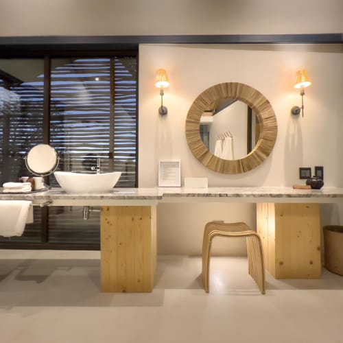 Ein modernes Hotelbadezimmer mit einer Marmor-Arbeitsplatte, zwei runden Waschtischen, einem Spiegel und wandmontierten Lampen, mit Blick auf einen Außenbereich durch Glas-Schiebetüren.