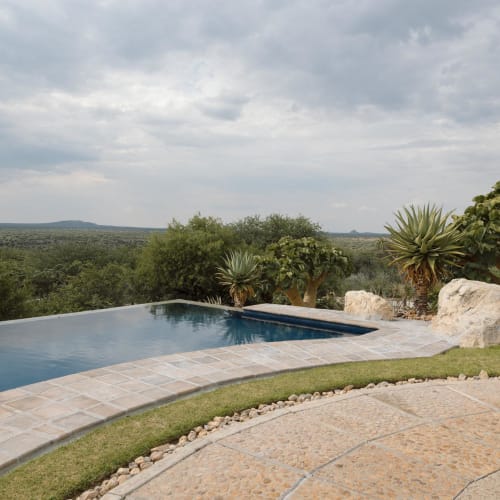 An infinity pool with a view of nature, surrounded by plants and stones, under a cloudy sky.