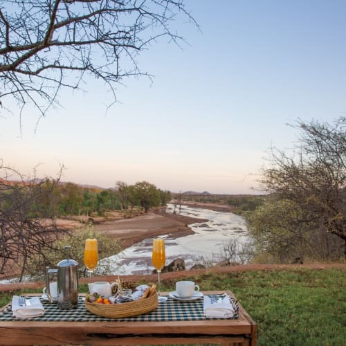 A breakfast setup on a wooden table overlooking a river with trees, in a savannah landscape during sunset.