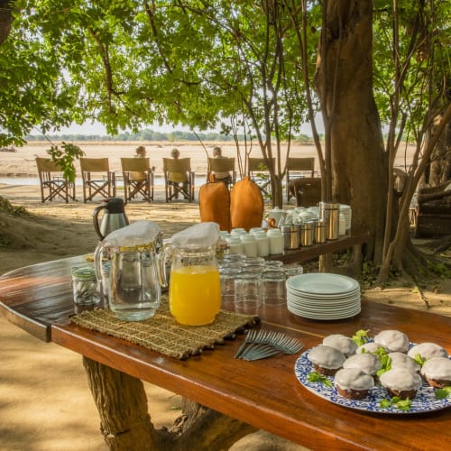 Breakfast setup on a wooden table under trees on a sandy beach, with chairs in the background.