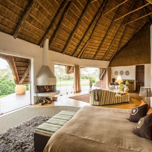 Geräumige Loungefläche in einer Lodge mit Strohdach, großen Fenstern und bequemer Einrichtung, darunter Bett, Sofa und Sessel.