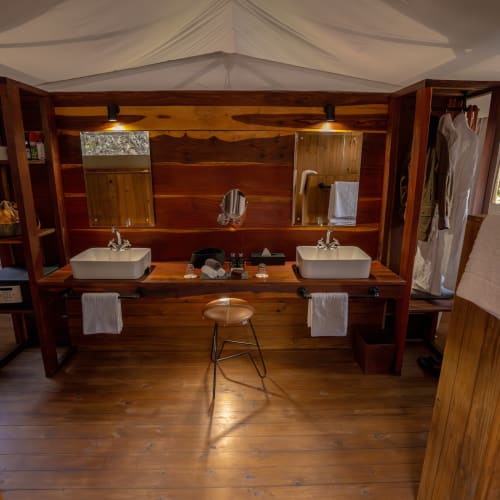 A rustic bathroom with double sinks, mirrors, and wooden walls, located in a nature setting with visible trees outside.