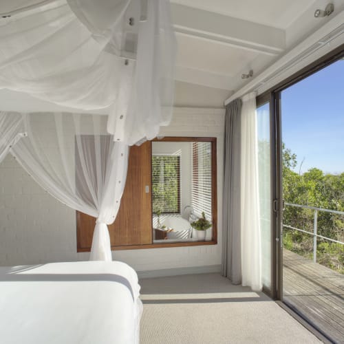 Helles Schlafzimmer mit weißem Himmelbett und großen, verglasten Schiebetüren, die auf eine Holzterrasse mit Blick auf grüne Bäume und einen klaren blauen Himmel hinausgehen.