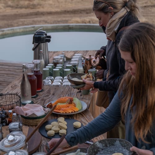 Zwei Frauen bedienen sich Frühstück an einem Outdoor-Tisch in der Nähe eines Sees bei Sonnenaufgang oder Sonnenuntergang, mit Bäumen und Bergen im Hintergrund.