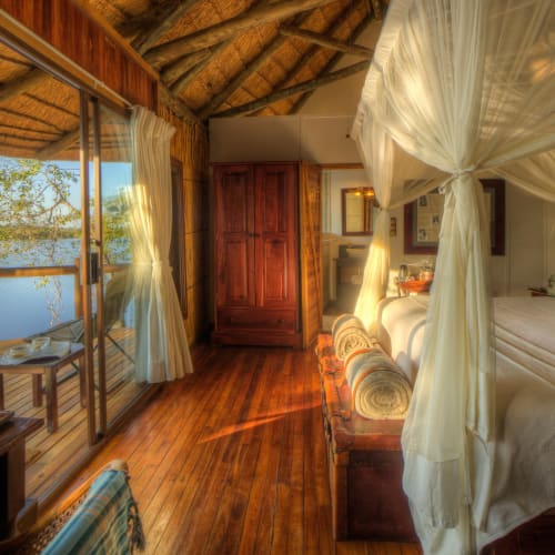 Interior of a luxurious lodge suite in Botswana with a large bed, mosquito netting, wooden furniture, and a view of the water through glass doors.