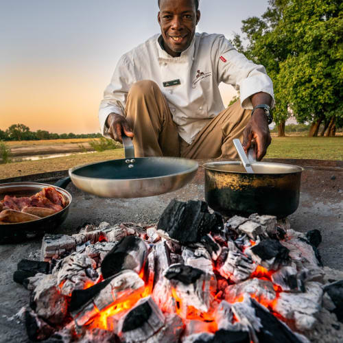 A chef grilling outdoors over a campfire with a sunset in the background