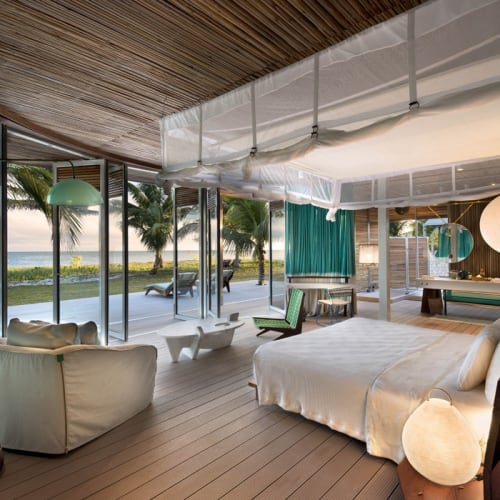 Luxurious bedroom with large windows overlooking a tropical beach with palm trees, furnished with a white bed, a sofa, and modern decor.