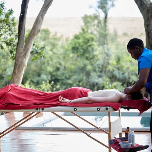 A person receiving a massage outdoors on a massage table draped with a red cloth, with a masseuse attending to them, surrounded by trees and greenery.