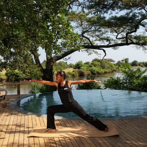 A woman practicing yoga beside an infinity pool with a scenic river and trees in Tanzania.