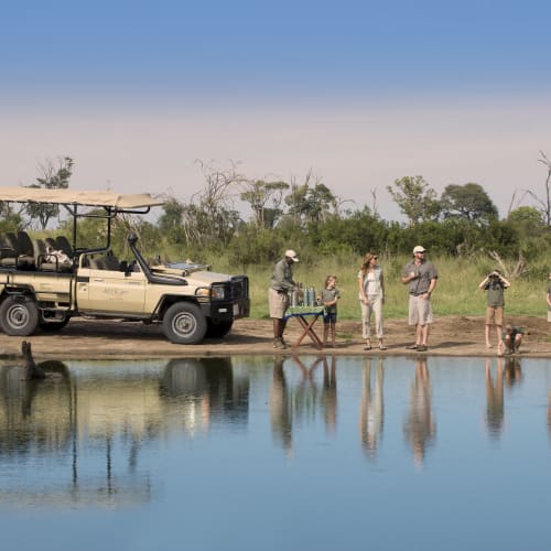 Touristen und Guides bereiten sich an einem Flussufer für ein Picknick vor, in der Nähe eines Safari-Fahrzeugs in einer Savannenlandschaft.