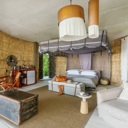 Luxuriöses Hotelzimmer mit einem Himmelbett, einem weißen Sessel, einem alten Koffer und Grünblick durch die Fenster.
