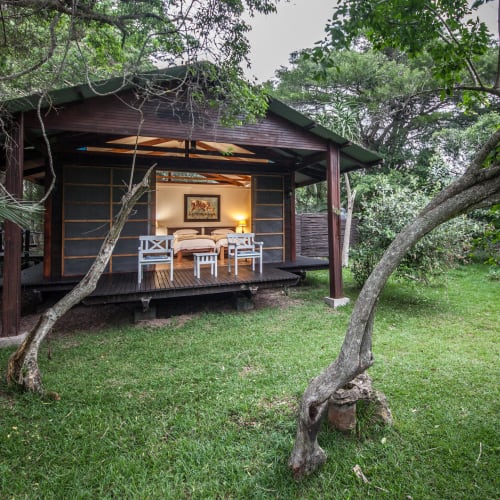 A cozy wooden lodge with an open front, situated among trees on a lush green lawn, showing a dining area inside.