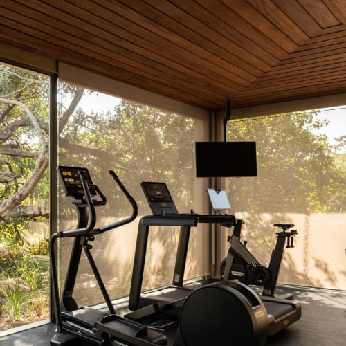 Modern fitness room with large windows showing outdoor greenery, containing an elliptical machine, a treadmill, a stationary bike, and a rowing machine.