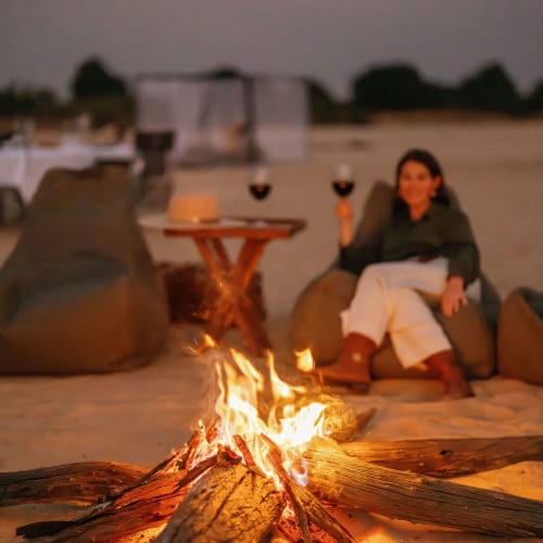 Eine Frau sitzt entspannt neben einem Strandfeuer bei Sonnenuntergang und hält ein Glas Rotwein.