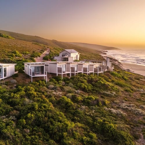 Moderne Gebäude auf einer Klippe mit Blick auf den Strand bei Sonnenuntergang, umgeben von grüner Vegetation.
