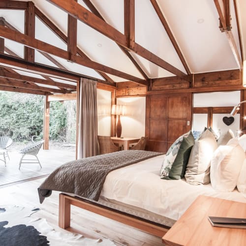 A cozy bedroom with wooden accents, large bed with pillows, and a sliding glass door opening to an outdoor seating area surrounded by greenery.