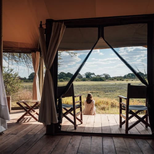 Eine Frau sitzt auf einer hölzernen Veranda vor einem Zelt und blickt auf eine weite Graslandschaft in Sambia.