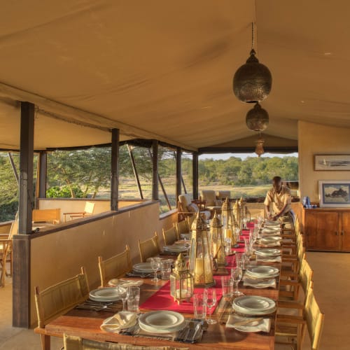 A spacious dining area with a long table set for a meal, surrounded by large windows overlooking greenery.