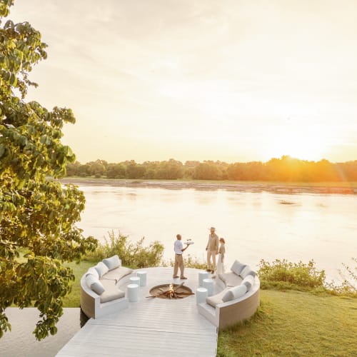 Zwei Personen stehen auf einem Holzdeck mit einer runden Sitzgelegenheit und Blick auf einen Fluss bei Sonnenuntergang, mit einem Lagerfeuer in der Mitte.