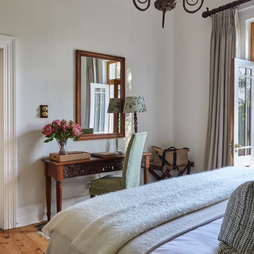 Bright bedroom with large windows, outdoor view, and vintage furniture including a bed, mirror, and desk with a chair.