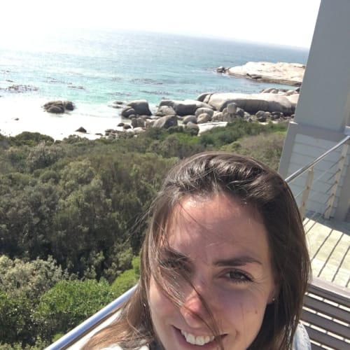 A smiling woman with brown hair taking a selfie on a balcony overlooking the ocean and rocks in South Africa.