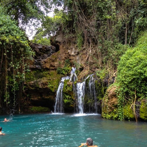 People swimming in a natural pool with a small waterfall surrounded by lush green vegetation in Kenya.