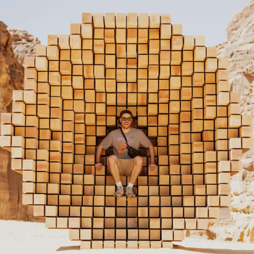 A man sitting inside a circular, honeycomb-like wooden structure set in a desert landscape with rocky cliffs in the background.